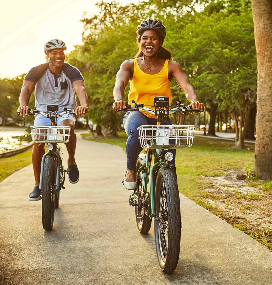 Two smiling riders on cruiser electric bikes with front baskets on a waterside path lined with palms